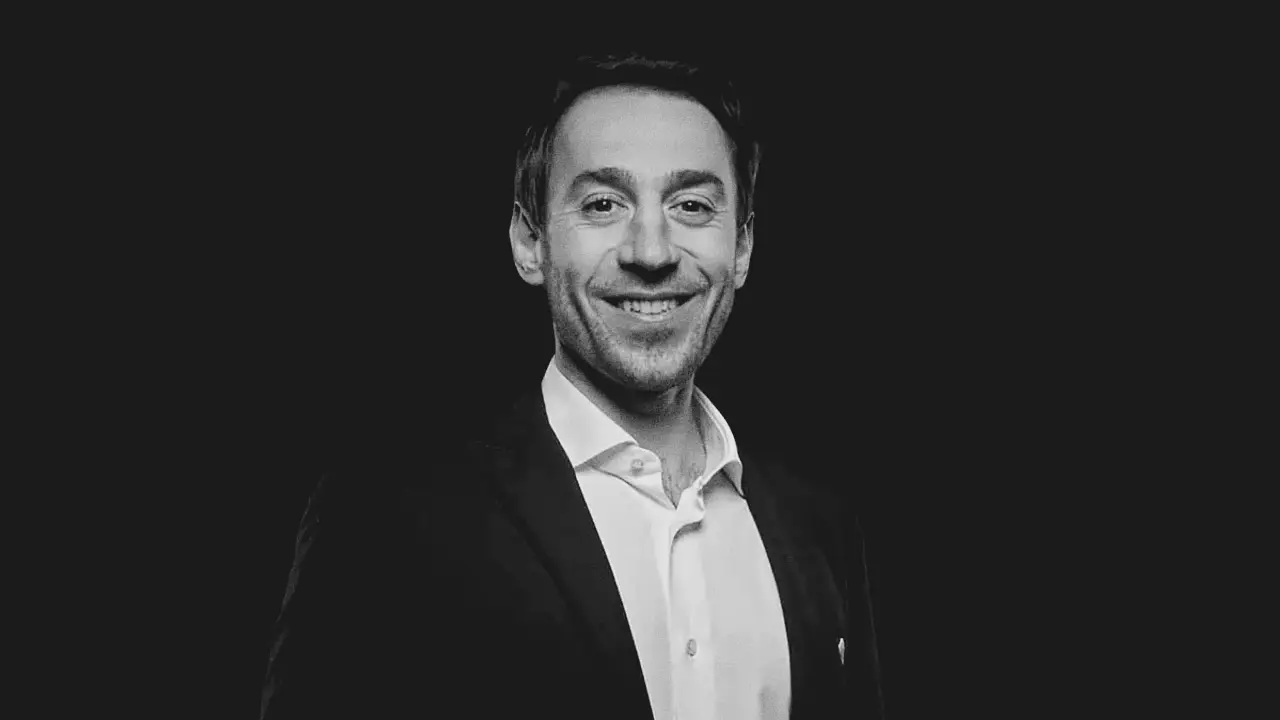 Uomo sorridente in giacca e camicia bianca, su sfondo scuro.