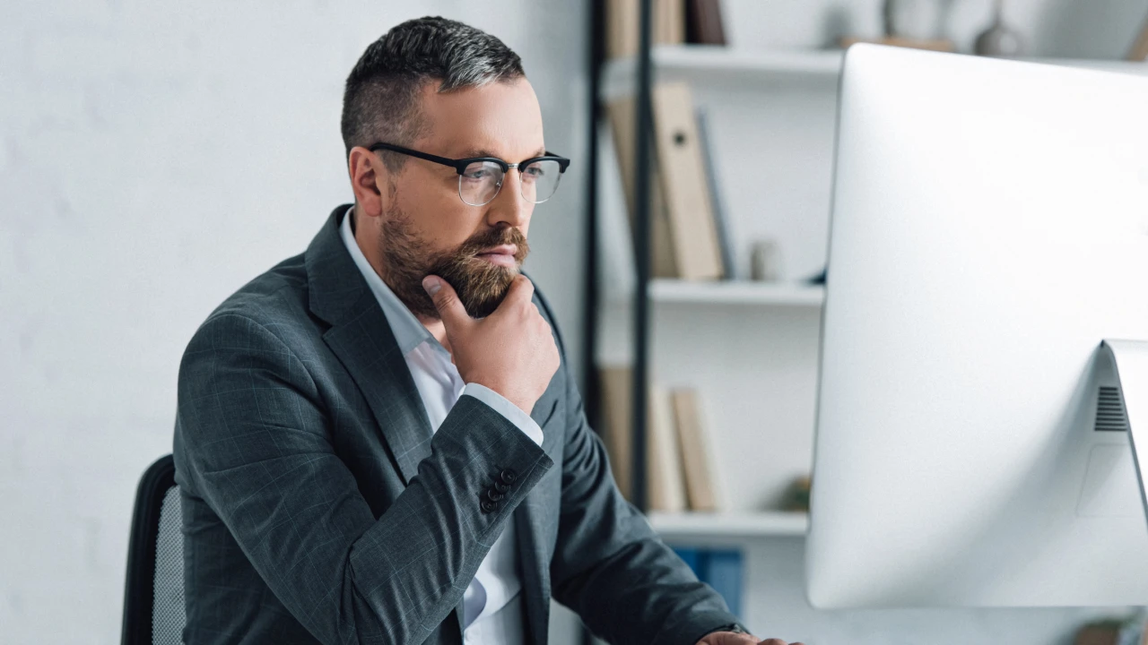 Uomo con barba e occhiali, in giacca e camicia, pensa davanti a un computer.
