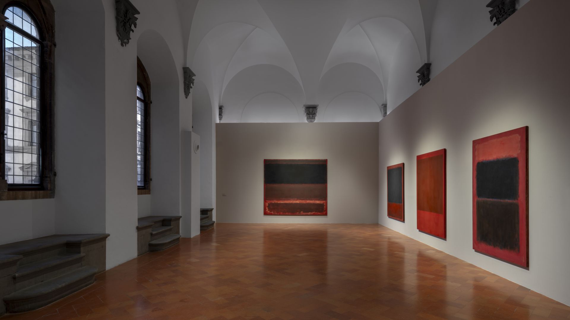 Galleria d'arte con dipinti astratti colorati su pareti bianche e pavimento in cotto.