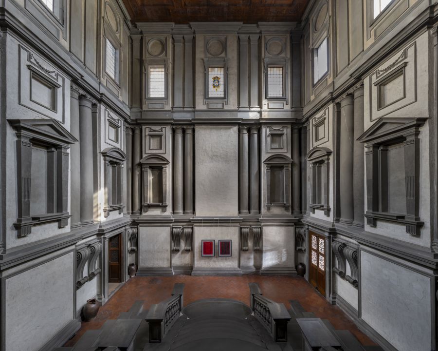 Interno di un edificio con pareti grigie, colonne, finestre e due quadri rossi.