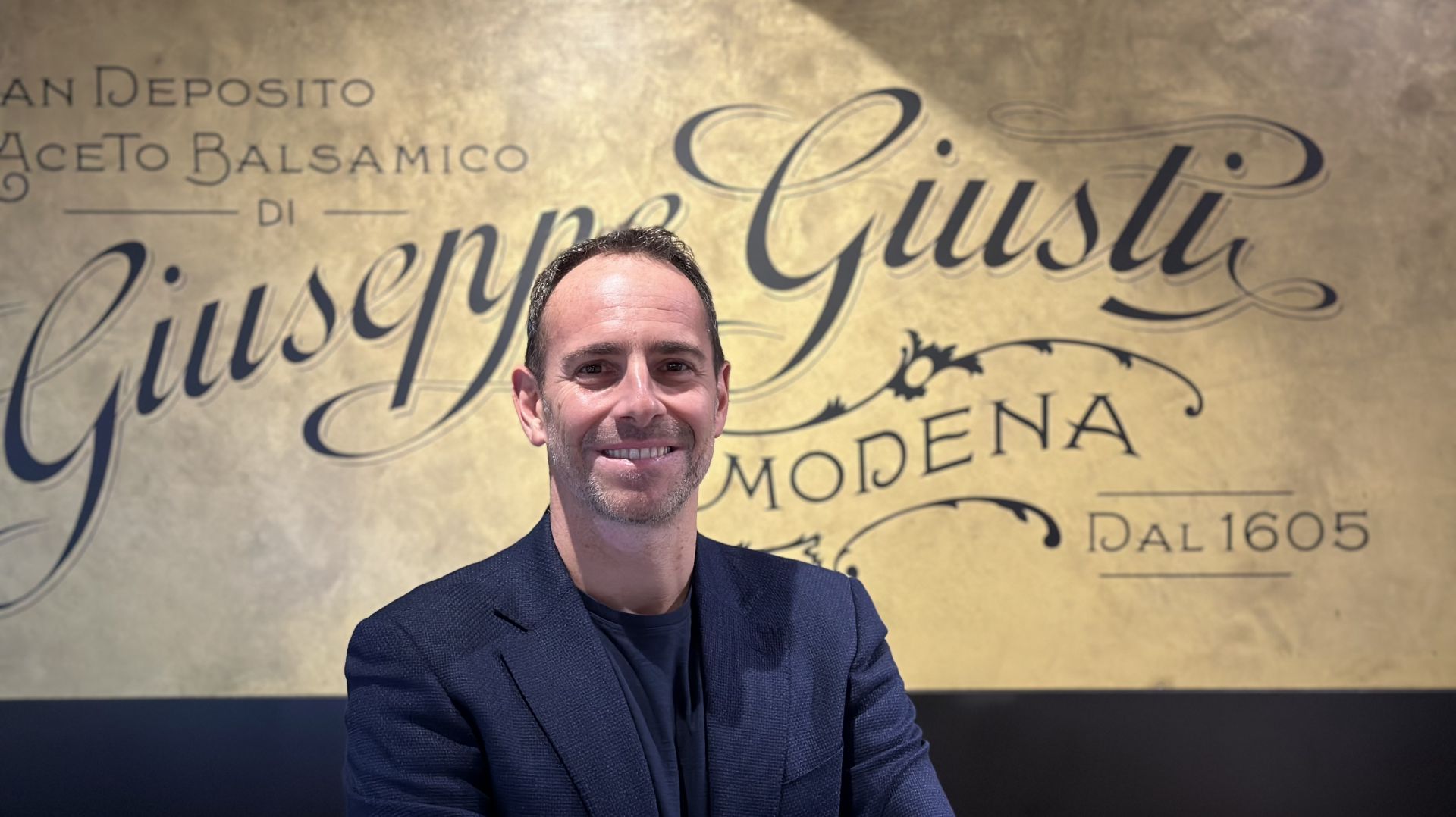 Un uomo sorridente in blazer scuro si trova di fronte a una parete dorata con un'elegante scritta "Giuseppe Giusti Modena dal 1605" e altre scritte in italiano relative all'aceto balsamico.