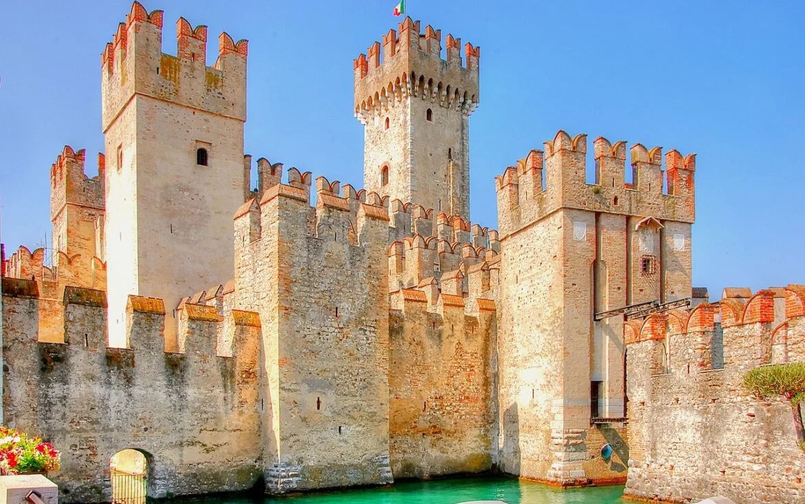 Un castello medievale in pietra con torri e mura merlate si trova accanto a un fossato turchese sotto un cielo azzurro e limpido. Una bandiera italiana sventola in cima alla torre centrale più alta: un'occasione ideale per chi desidera comprare castello in Italia.