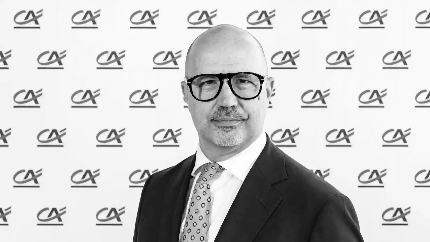 Uomo con occhiali e cravatta a pois, sfondo con logo ripetuto.