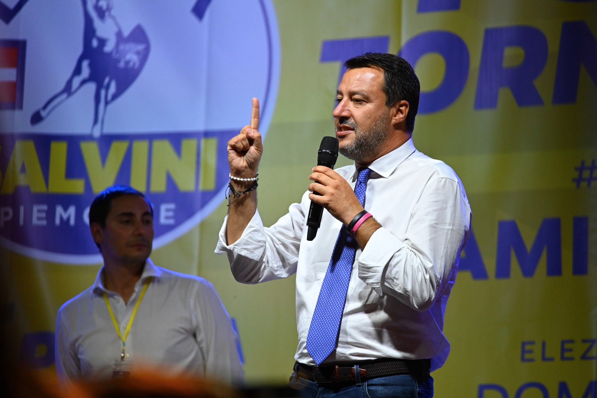 Uomo con camicia bianca e cravatta blu parla al microfono, indice alzato.