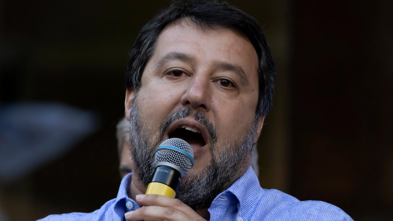 Uomo con barba grigia parla al microfono, indossa camicia azzurra a righe.