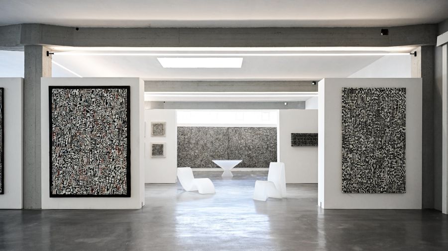 Galleria d'arte moderna con opere astratte, sedie bianche e un tavolo scultoreo.
