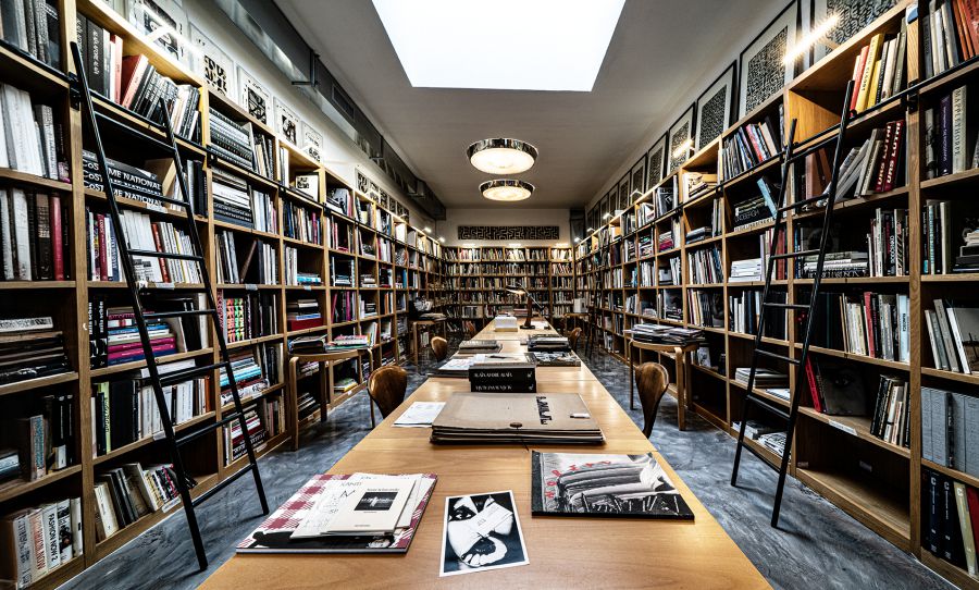 Libreria con scaffali pieni di libri, tavoli lunghi e sedie in legno.