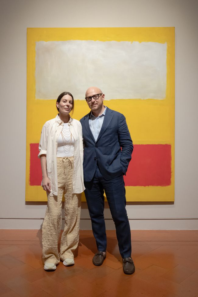 Coppia posa davanti a un dipinto astratto con blocchi di colore giallo, bianco e rosso.