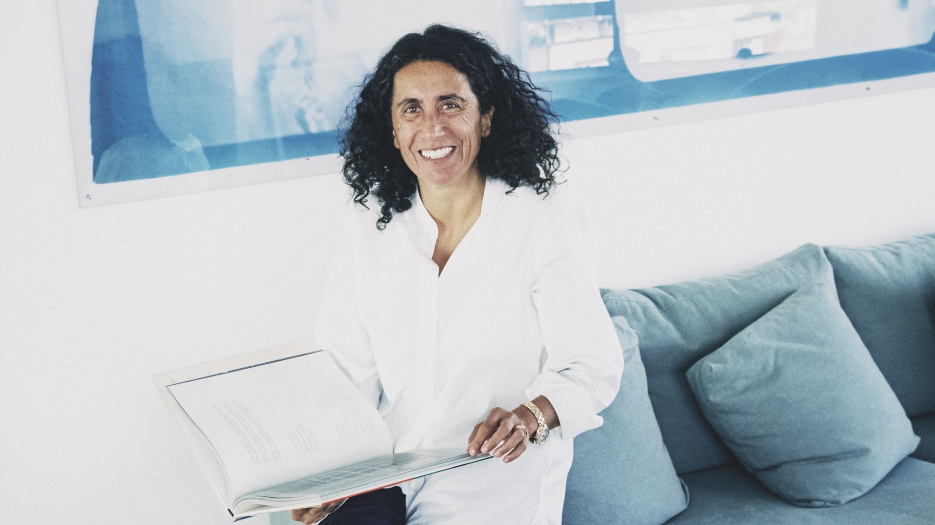 Donna sorridente con capelli ricci scuri, tiene un libro aperto, seduta su un divano blu.