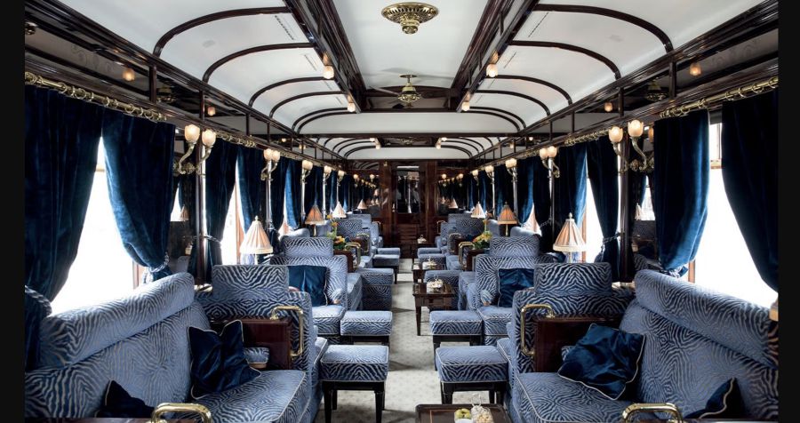 Interni lussuosi di carrozze ferroviarie che ricordano l'Orient Express di oggi, con sedili in peluche con motivi blu, pannelli in legno scuro, accenti dorati, piccole lampade da tavolo e ampie finestre drappeggiate con ricche tende blu per un'elegante atmosfera vintage.