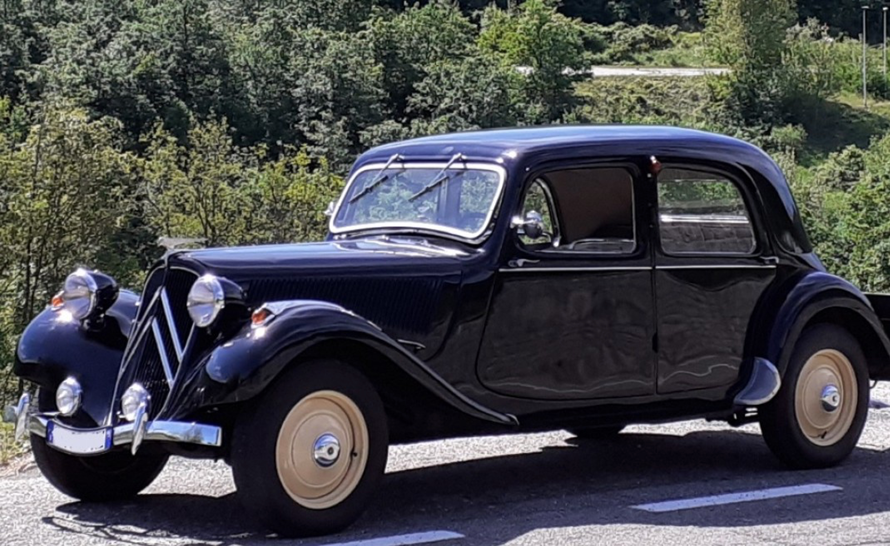 Auto d'epoca nera con parafanghi curvi, fari rotondi e ruote color crema, su strada asfaltata con sfondo verde.
