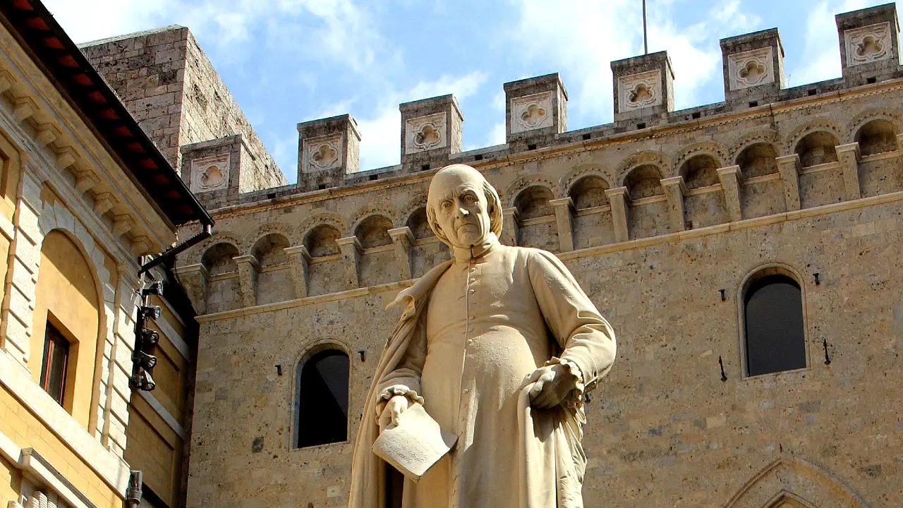 Statua di uomo con toga e foglio, davanti a palazzo storico con merlature.