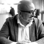 Uomo calvo con barba, occhiali, giacca e camicia bianca, mani giunte.