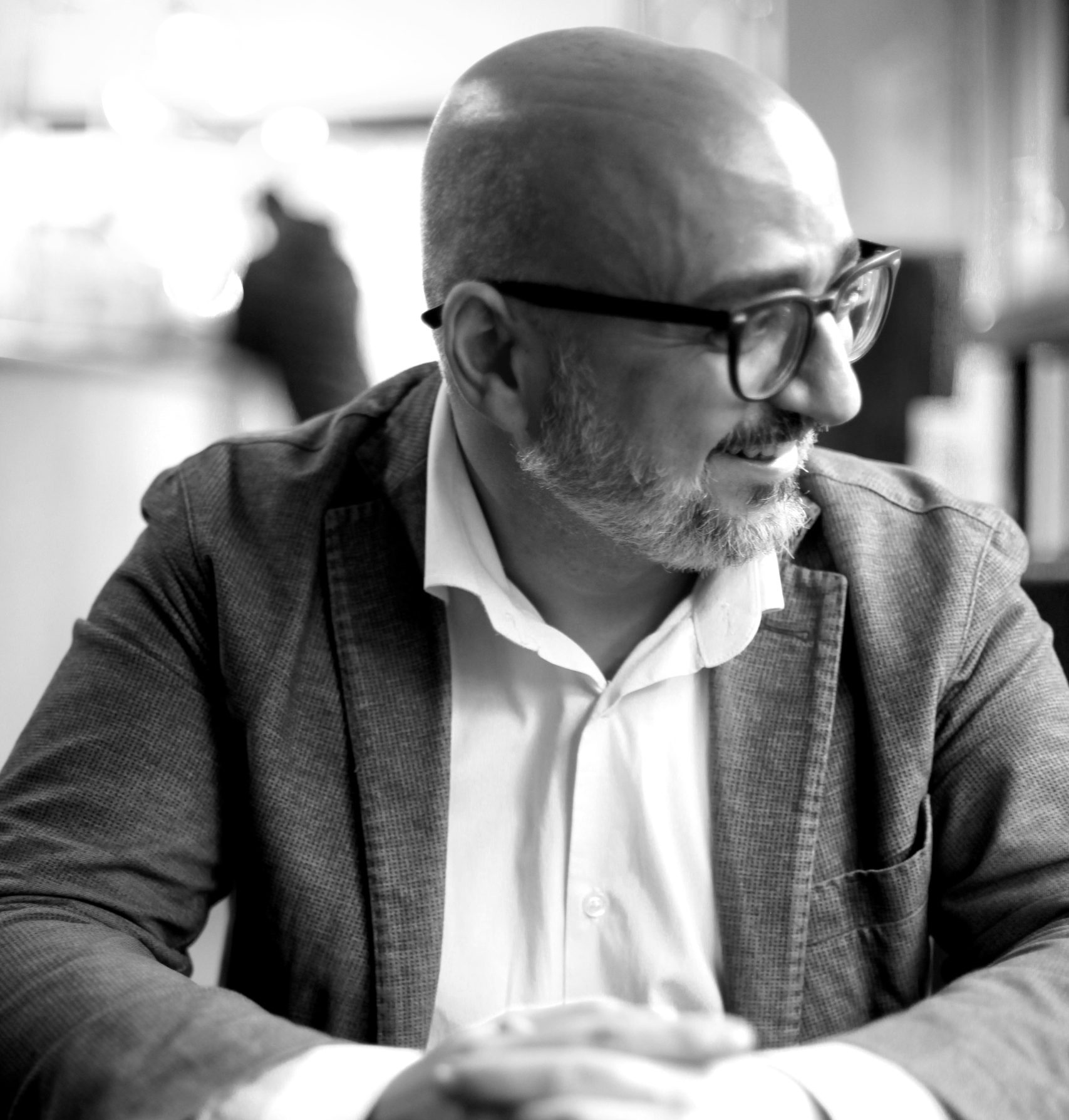 Uomo calvo con barba, occhiali, giacca e camicia bianca, mani giunte.