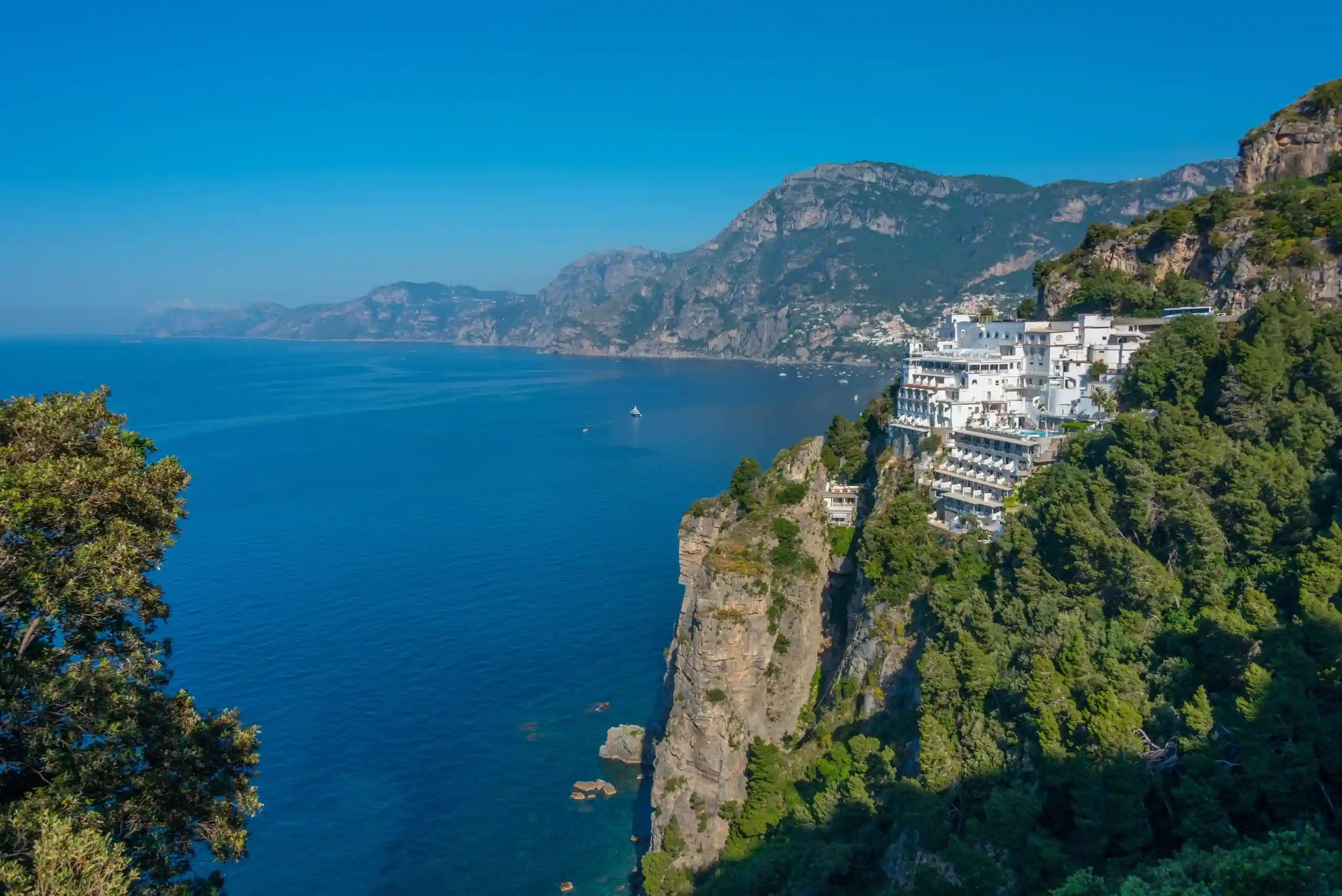 Edificio bianco su scogliera verde con mare blu e montagne sullo sfondo.