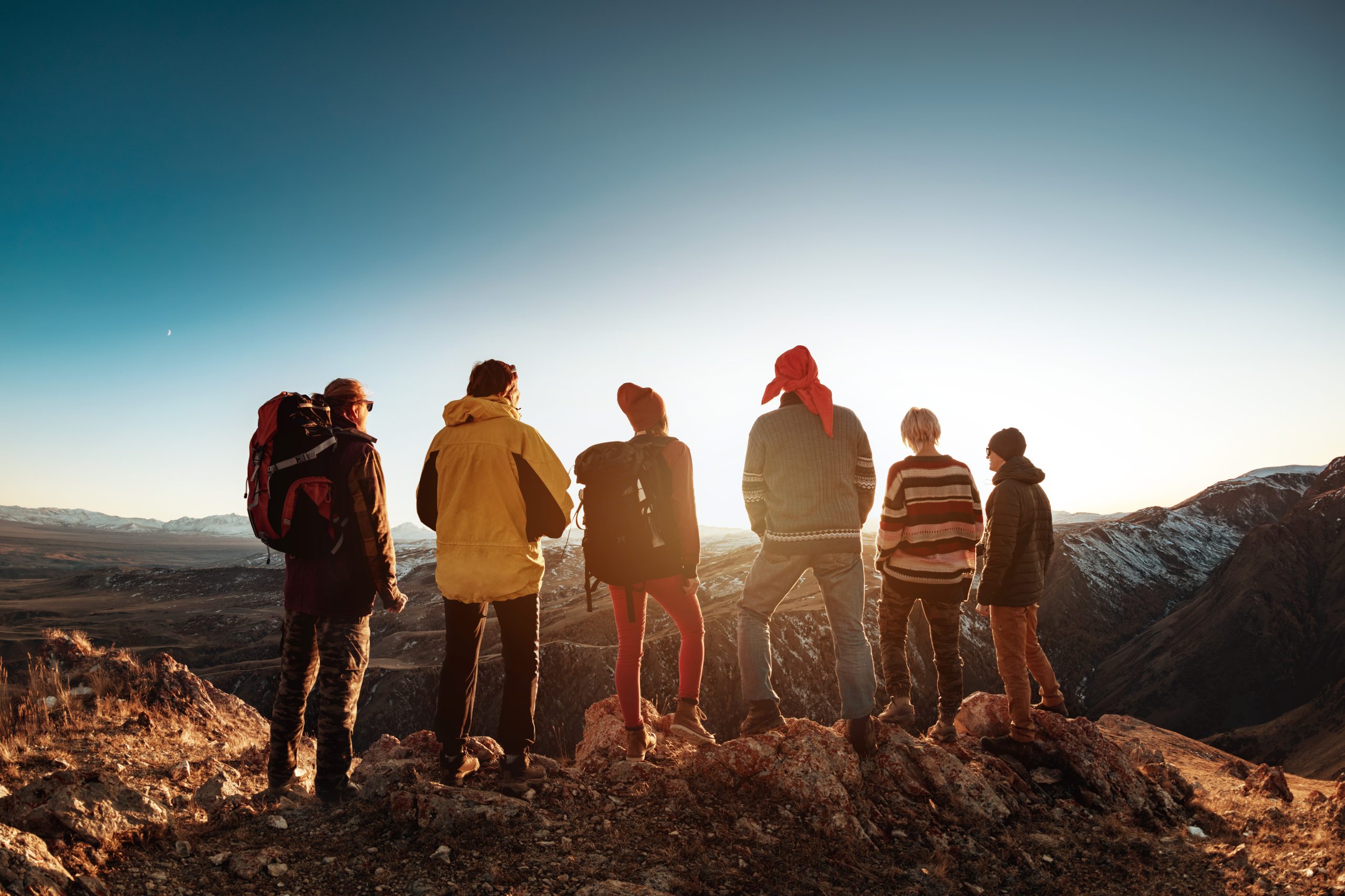 Sei persone in piedi su una cresta rocciosa guardano un vasto paesaggio montano al tramonto.