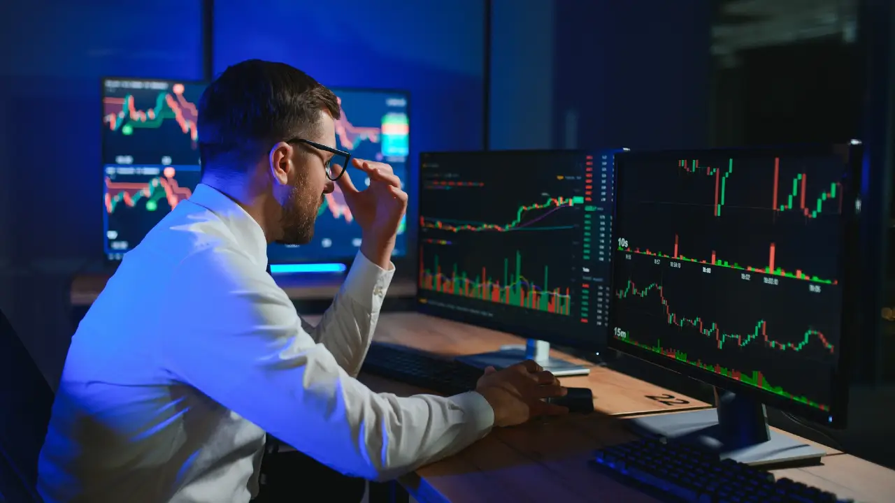 Uomo con occhiali guarda grafici finanziari su più monitor in una stanza buia illuminata da luci blu.