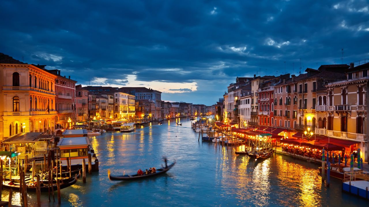 Canale di Venezia illuminato di notte, con gondole e palazzi storici.