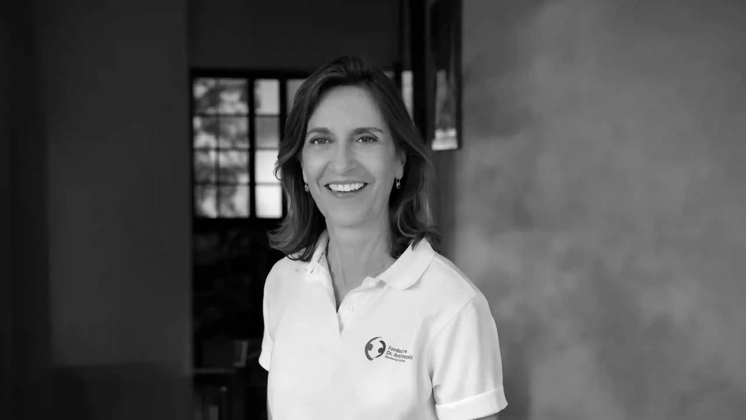 Donna sorridente con polo bianca con logo, sfondo sfocato con finestra.