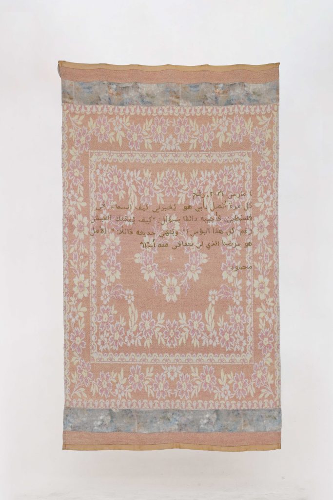 Tessuto rosa con motivi floreali bianchi e gialli, bordi decorati e testo arabo dorato.