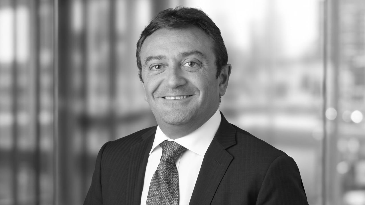 Uomo sorridente in abito scuro e cravatta con motivo geometrico, sfondo sfocato.