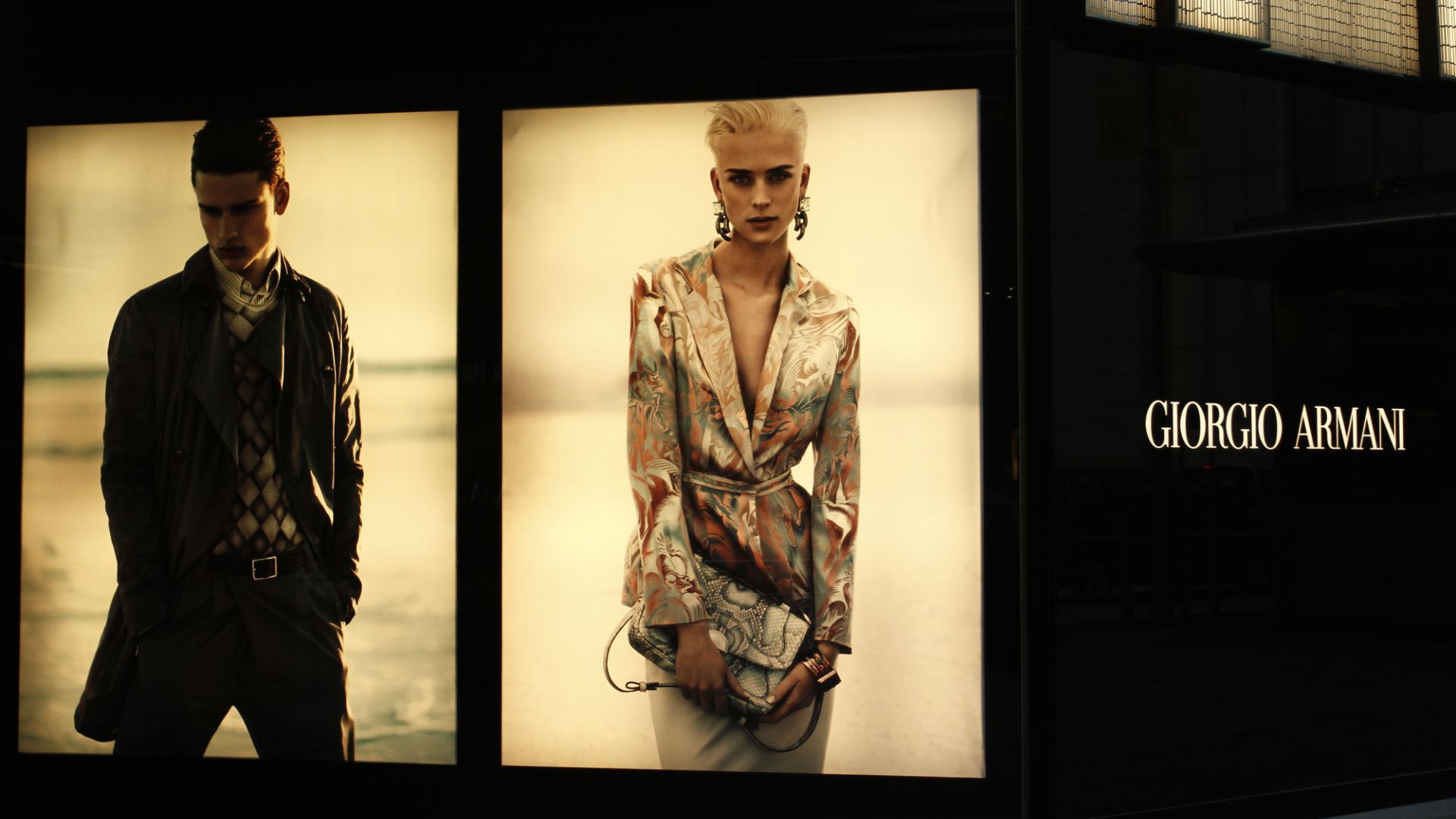 Due pubblicità di moda mostrano un uomo in giacca e sciarpa con lo sguardo rivolto verso il basso e una donna in abito a fantasia con in mano una pochette, catturando l'essenza della moda dazi. Il logo Giorgio Armani appare a destra su sfondo nero.