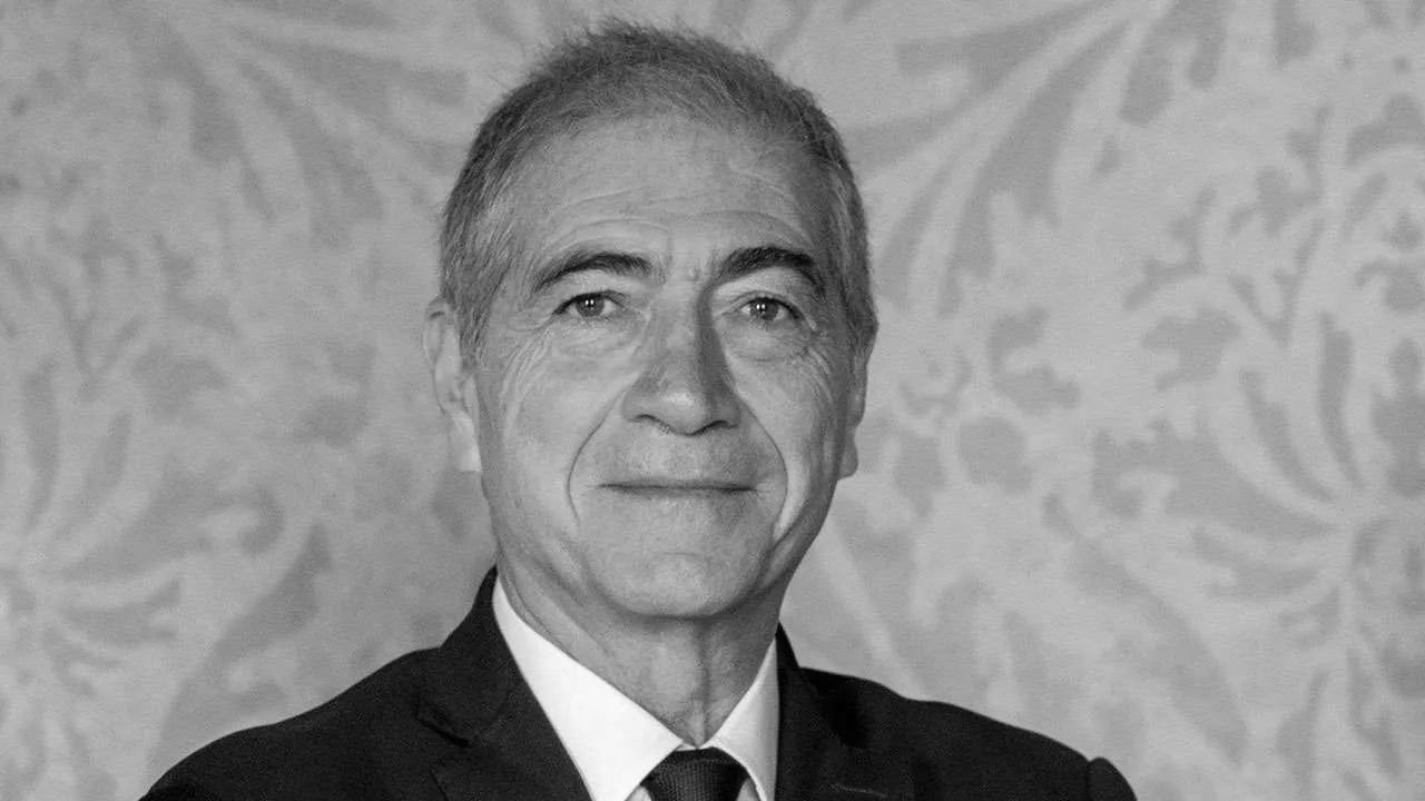 Ritratto in bianco e nero di un uomo anziano con abito scuro e cravatta, su sfondo decorato.