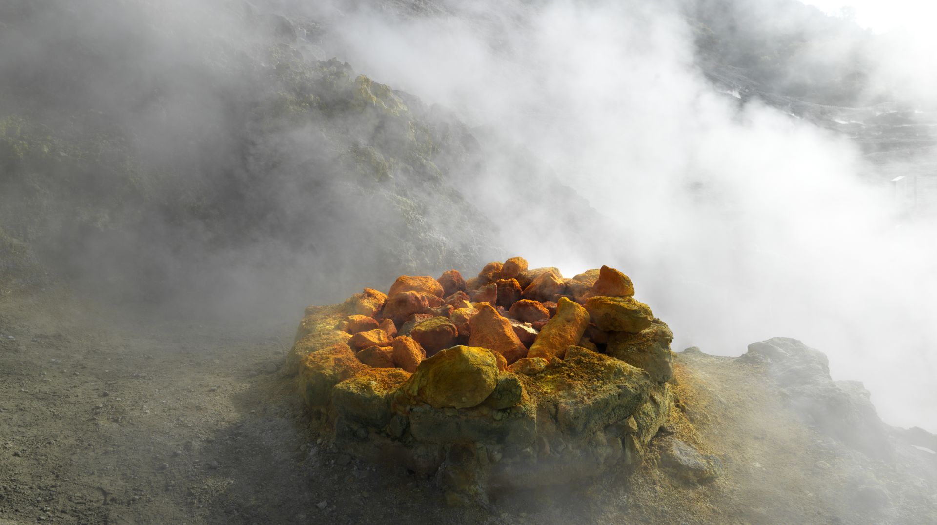 Rocce di zolfo giallo e arancione, raccolte in un mucchio circolare, emettono vapore, circondate da un paesaggio vulcanico roccioso e nebbioso: una scena ultraterrena dell'area di Panorama Pozzuoli.