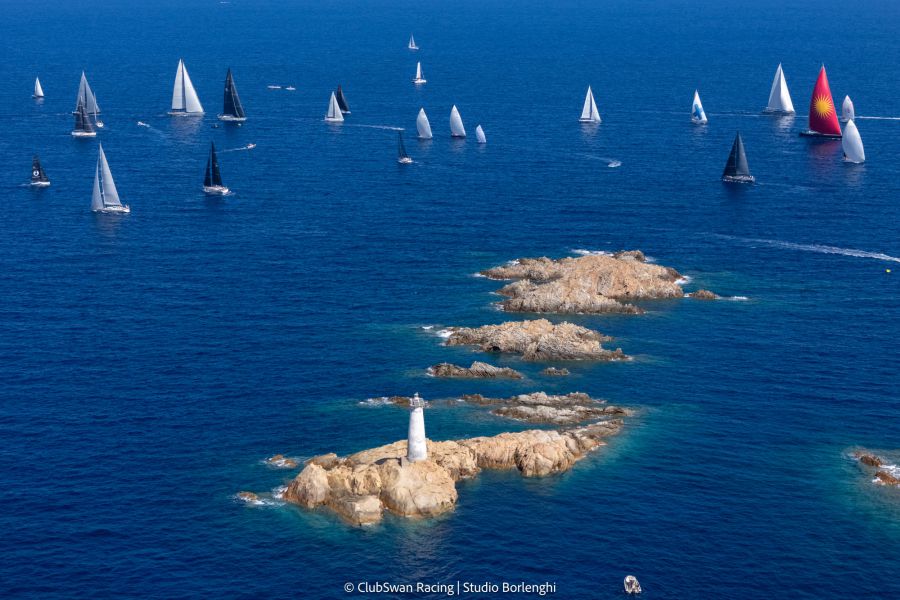 Barche a vela bianche e nere navigano in mare blu vicino a isole rocciose con un faro bianco.
