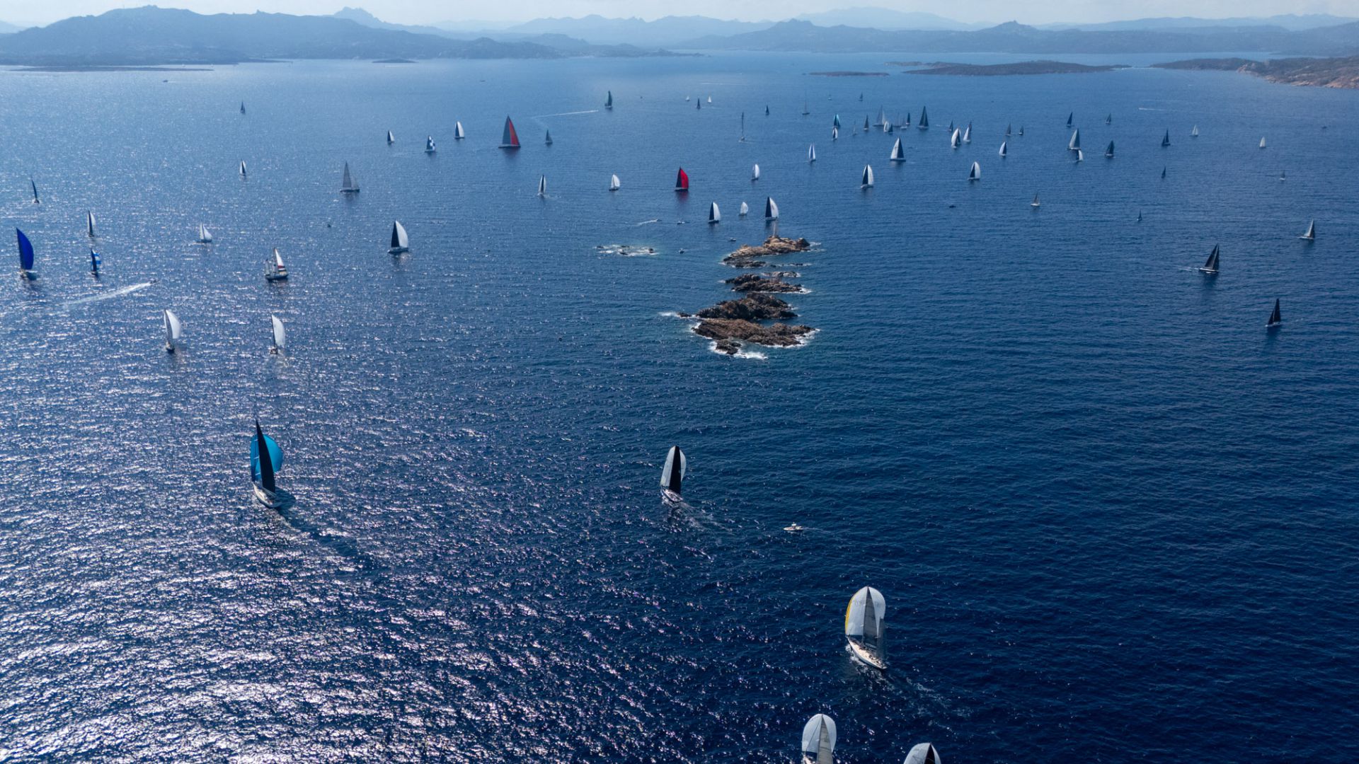 Regata di barche a vela con vele bianche, blu e rosse su mare blu intenso.