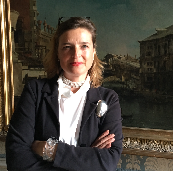 Donna con giacca scura, perle e bracciale, davanti a un dipinto di Venezia.