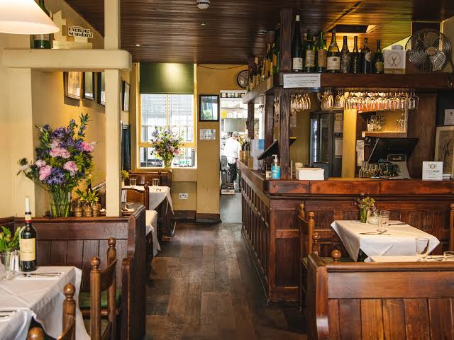 Bottiglia di vino, fiori, tavoli apparecchiati e bancone bar in legno scuro.