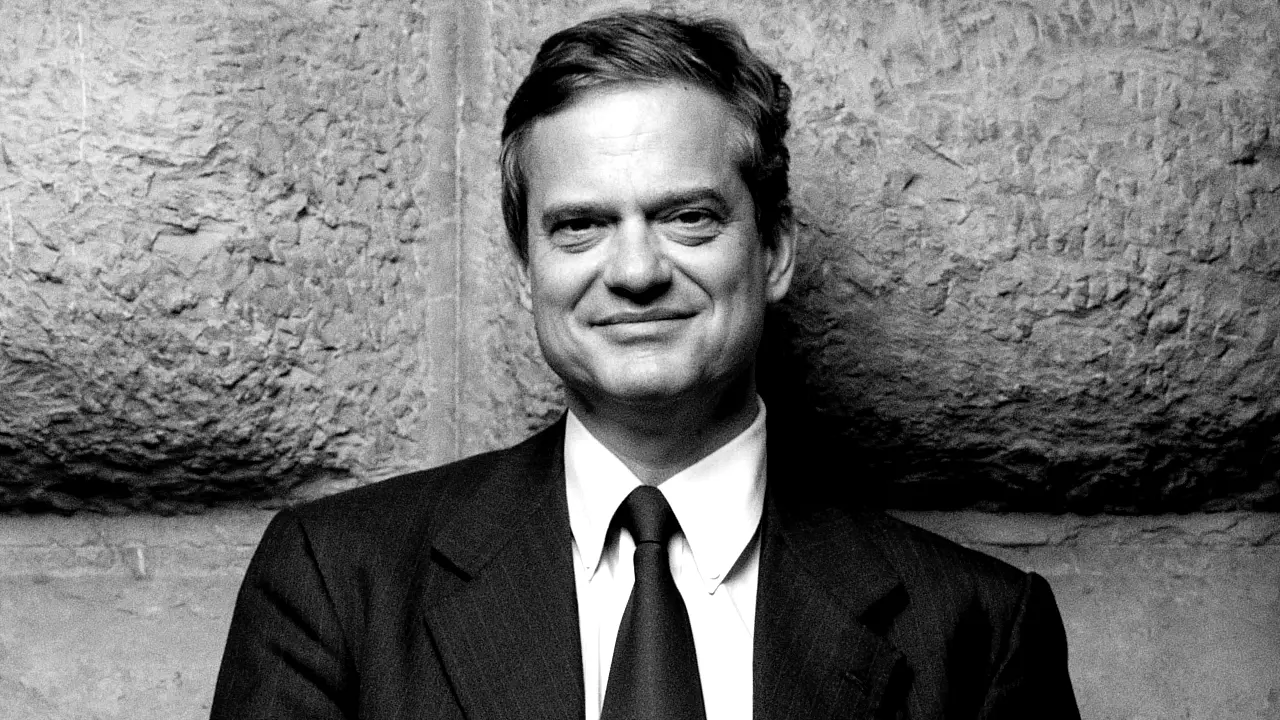 Uomo sorridente in abito scuro e cravatta, sfondo in pietra grezza.