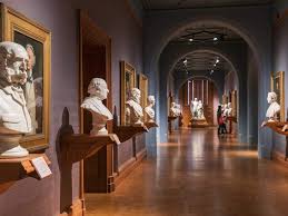 Busti in marmo e dipinti adornano le pareti di un corridoio museale con persone che passeggiano.