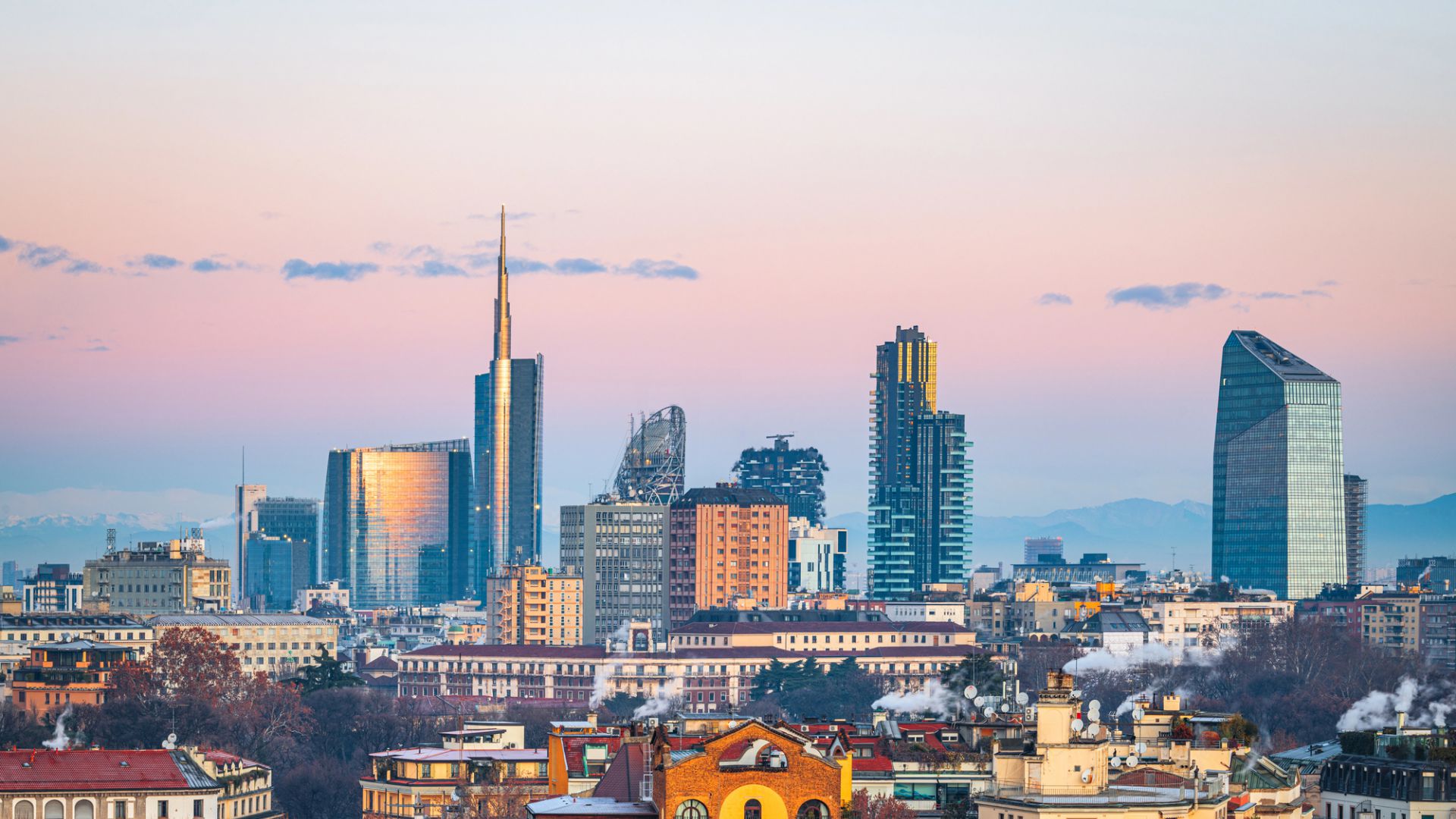 Skyline di grattacieli moderni e antichi edifici con montagne sullo sfondo al tramonto.
