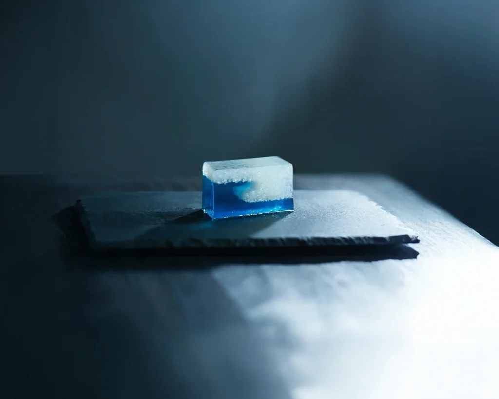 Cubetto di ghiaccio blu e bianco su una lastra scura, con sfondo sfocato.