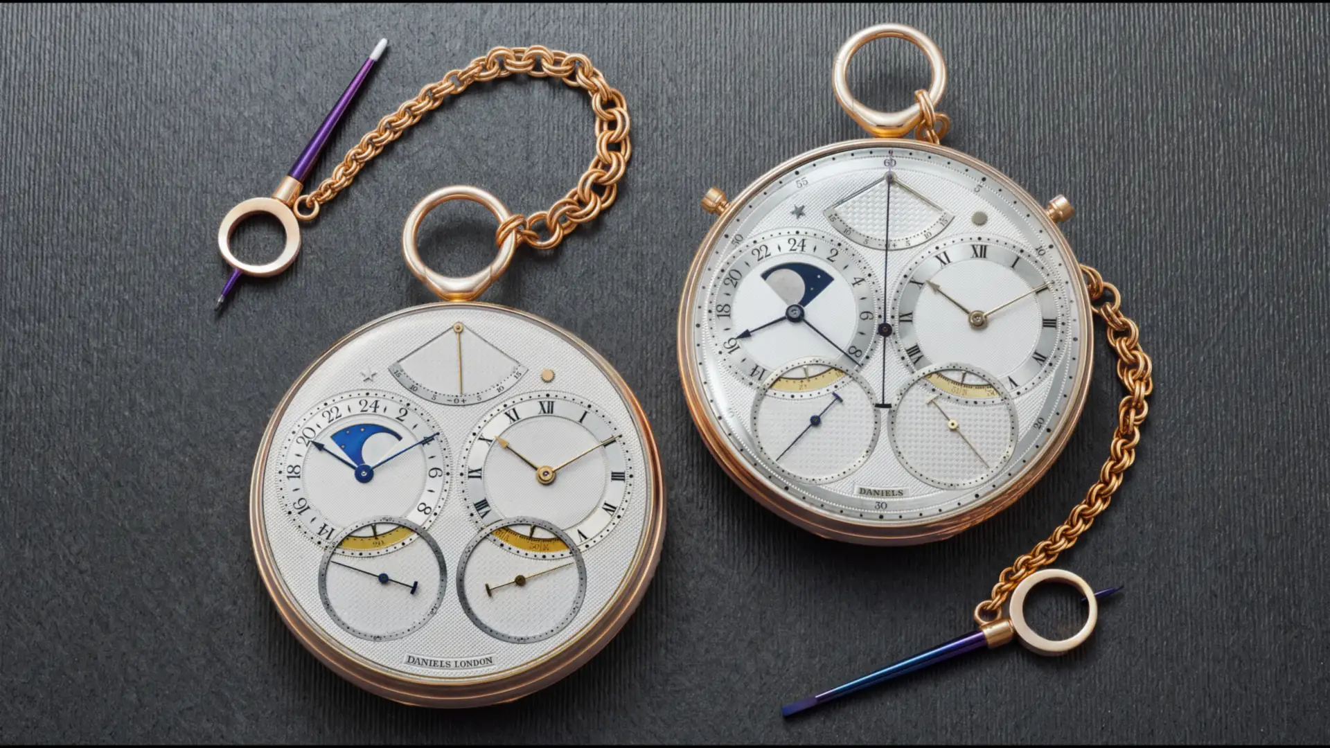 Due orologi da taschino dorati con quadranti complessi e catene, su sfondo scuro.