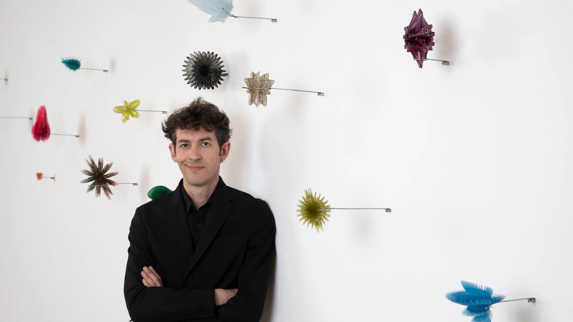 Uomo in giacca nera con braccia incrociate, appoggiato a un muro bianco con installazioni artistiche colorate.