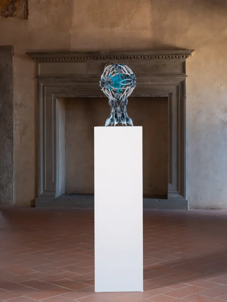 Scultura astratta con globo blu scintillante, su piedistallo bianco, davanti a un camino antico.