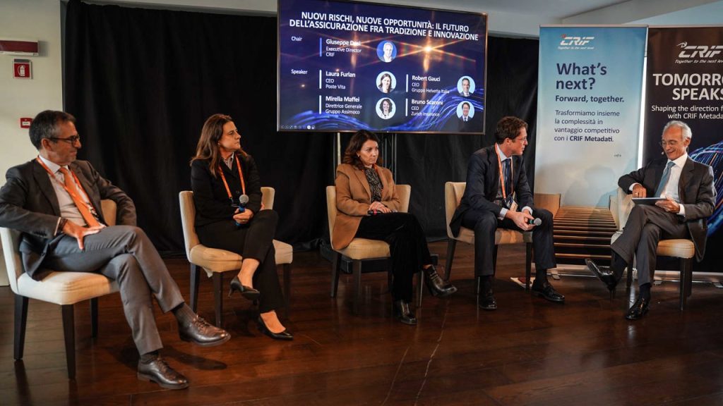 Panel di relatori seduti su sedie, con schermo retrostante che mostra nomi e titoli.