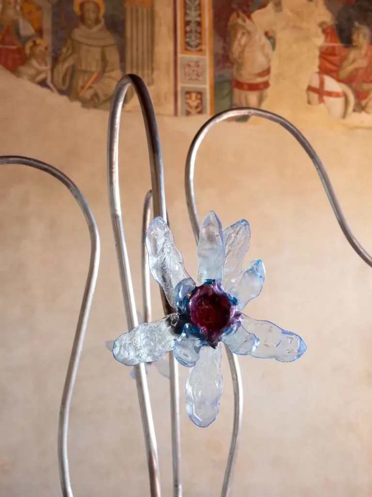 Fiore di vetro azzurro con centro viola scuro, su stelo metallico curvo, con affresco sullo sfondo.