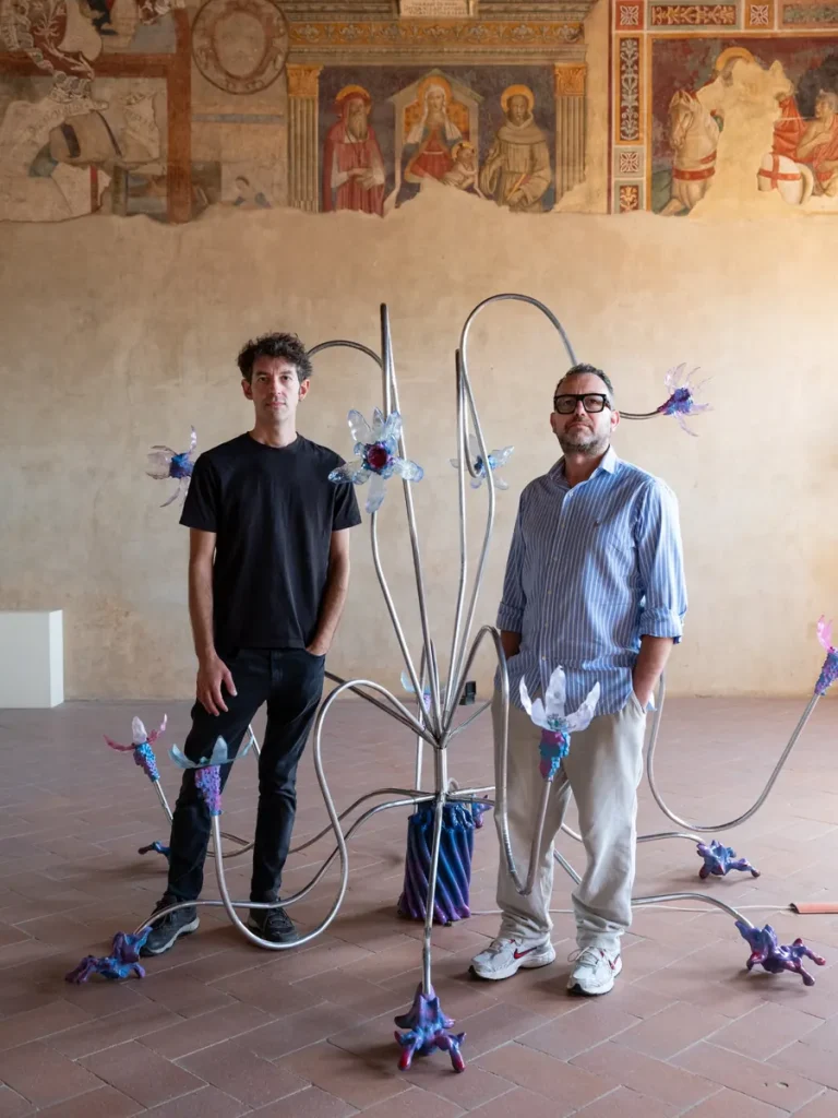 Due uomini posano accanto a un'installazione artistica metallica con elementi colorati simili a fiori.