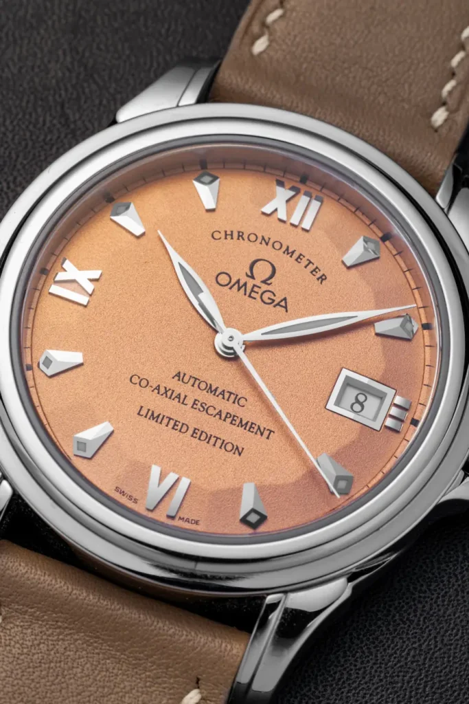 Orologio Omega con quadrante color salmone, indici romani e datario. Cinturino in pelle marrone.