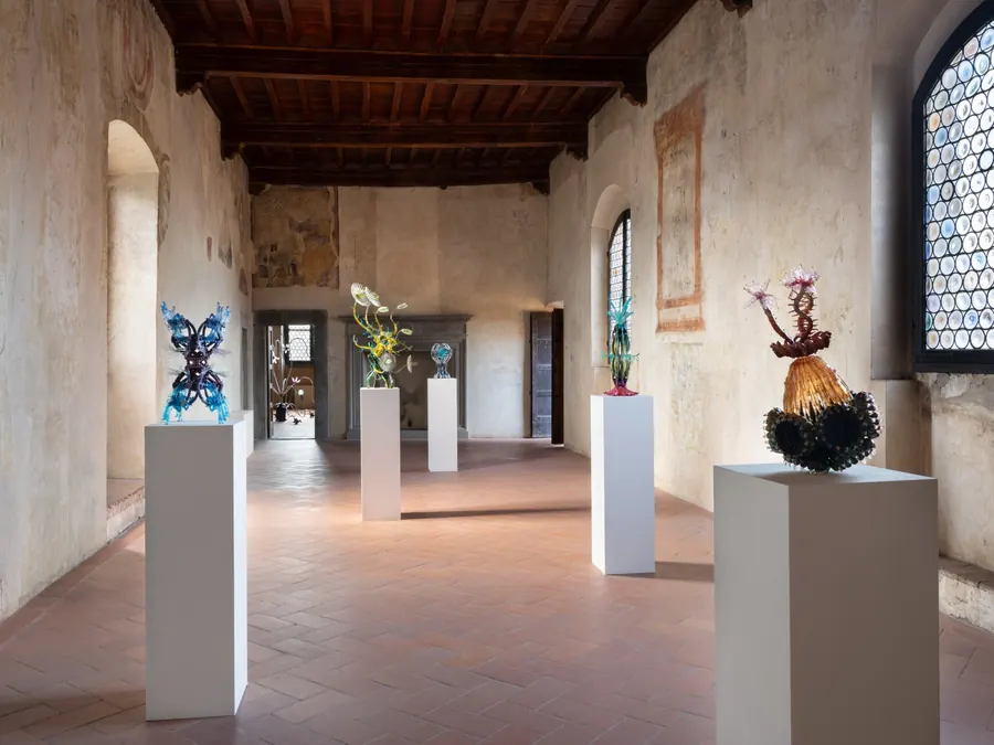 Sculture colorate di vetro su piedistalli bianchi in una sala con affreschi e finestre a grata.