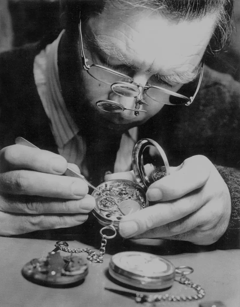 Orologiaio con lente d'ingrandimento ripara un orologio da tasca aperto, altri orologi sul tavolo.