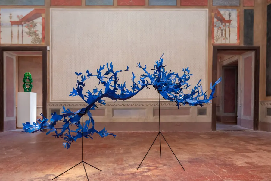 Scultura blu a forma di ramo su treppiedi, con una scultura verde su piedistallo a sinistra.