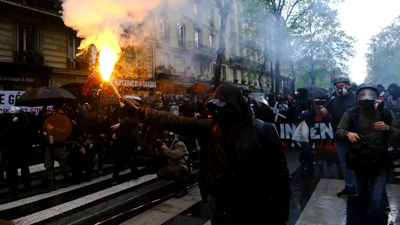 Manifestanti con fumogeni e maschere in strada, con edifici sullo sfondo.