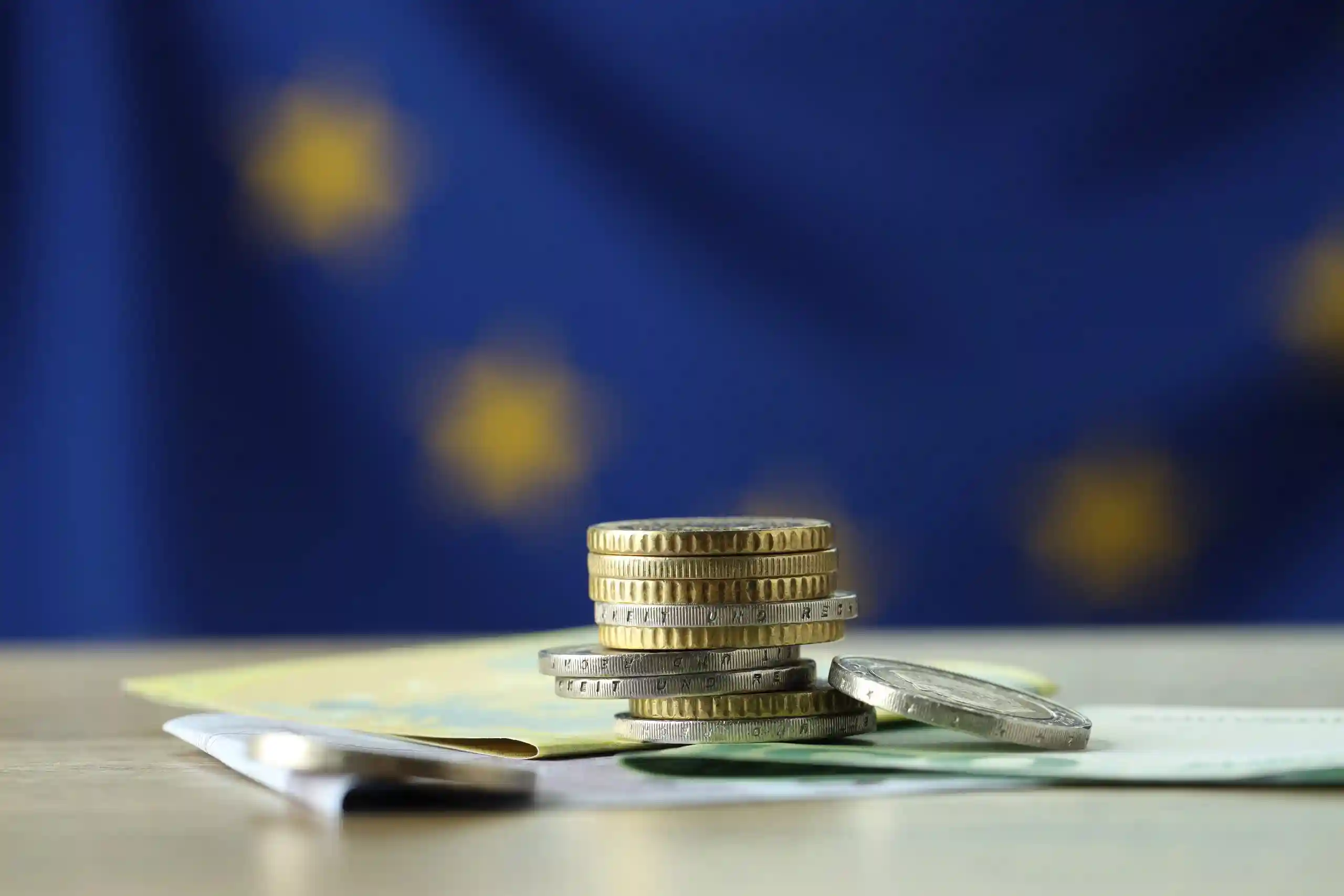Monete impilate su banconote con bandiera europea sfocata sullo sfondo.