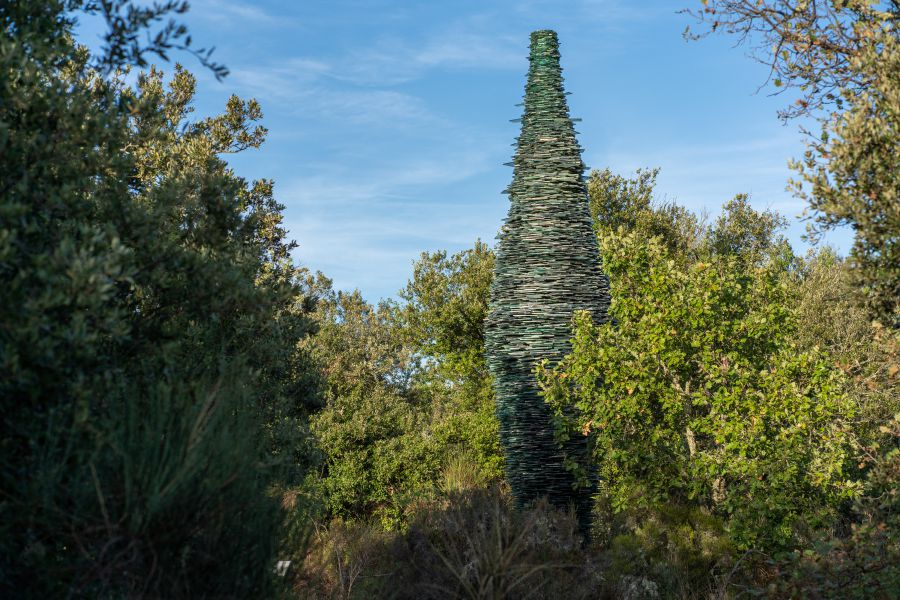 Scultura conica verde scuro fatta di strati, circondata da alberi e cielo blu.