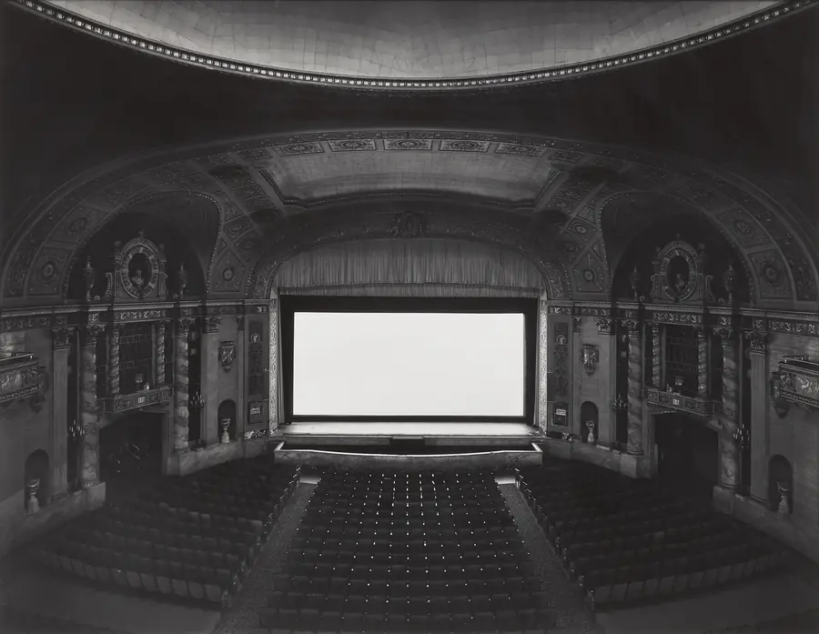 Sala cinematografica vuota con schermo bianco luminoso e sedili scuri.